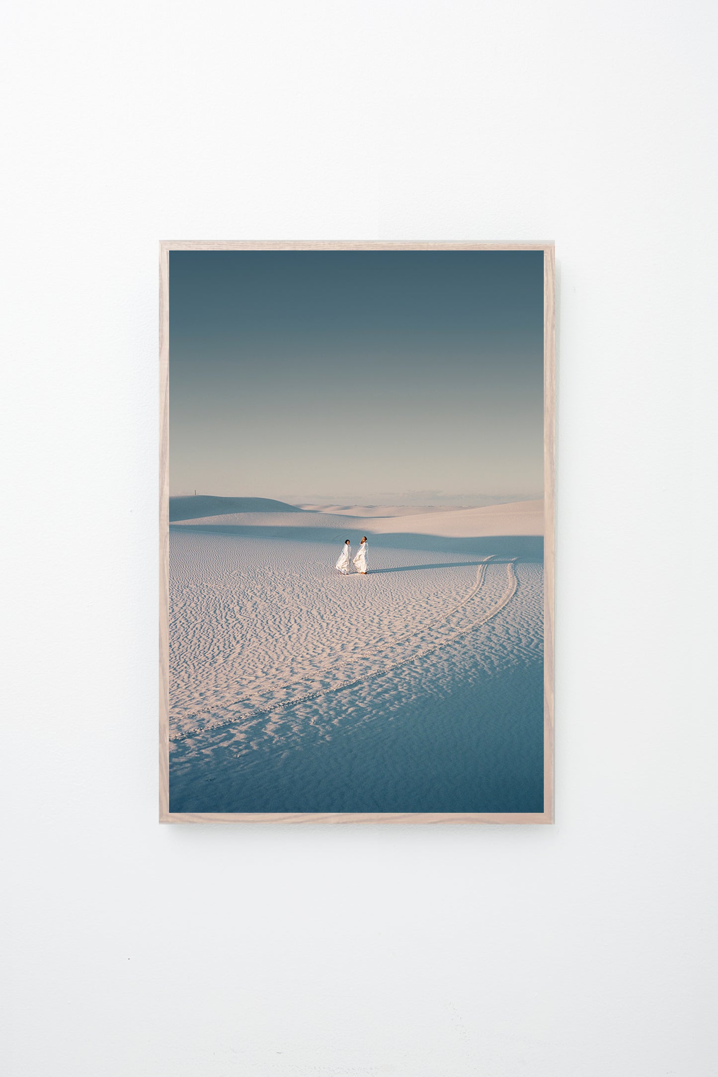 Two people stand in a desert scape, tire tracks beside them, framed on a white wall.