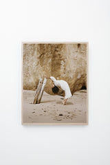 Woman leans backward and grabs onto driftwood, framed on white wall.