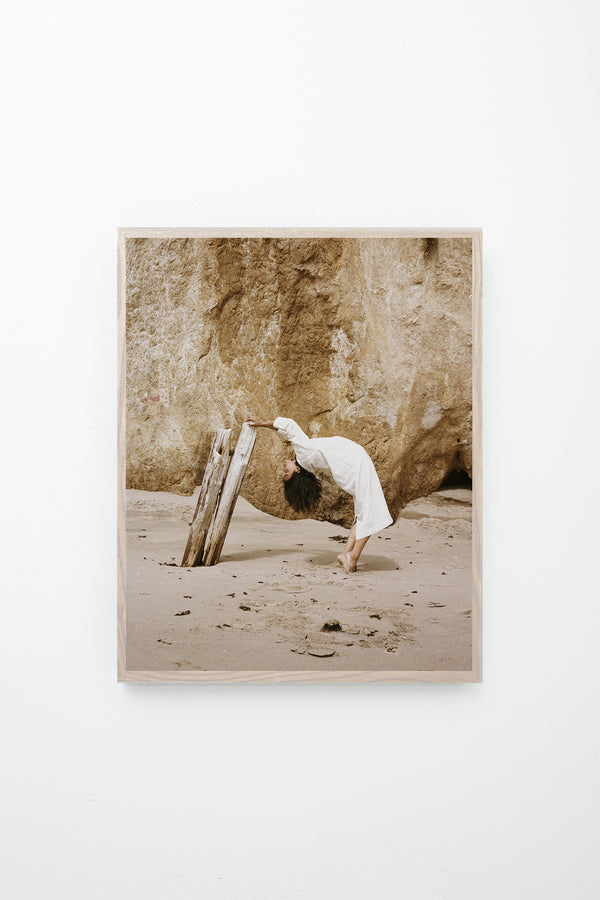 Woman leans backward and grabs onto driftwood, framed on white wall.